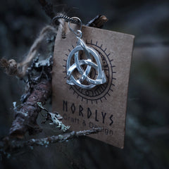 En silverbelagd evighetsknut-hängsmycke med trekantigt vävmönster, upphängd från en metallslinga framför en NORDLYS-märkt kraftpappersask med sollogga, omgiven av grenar och mossa