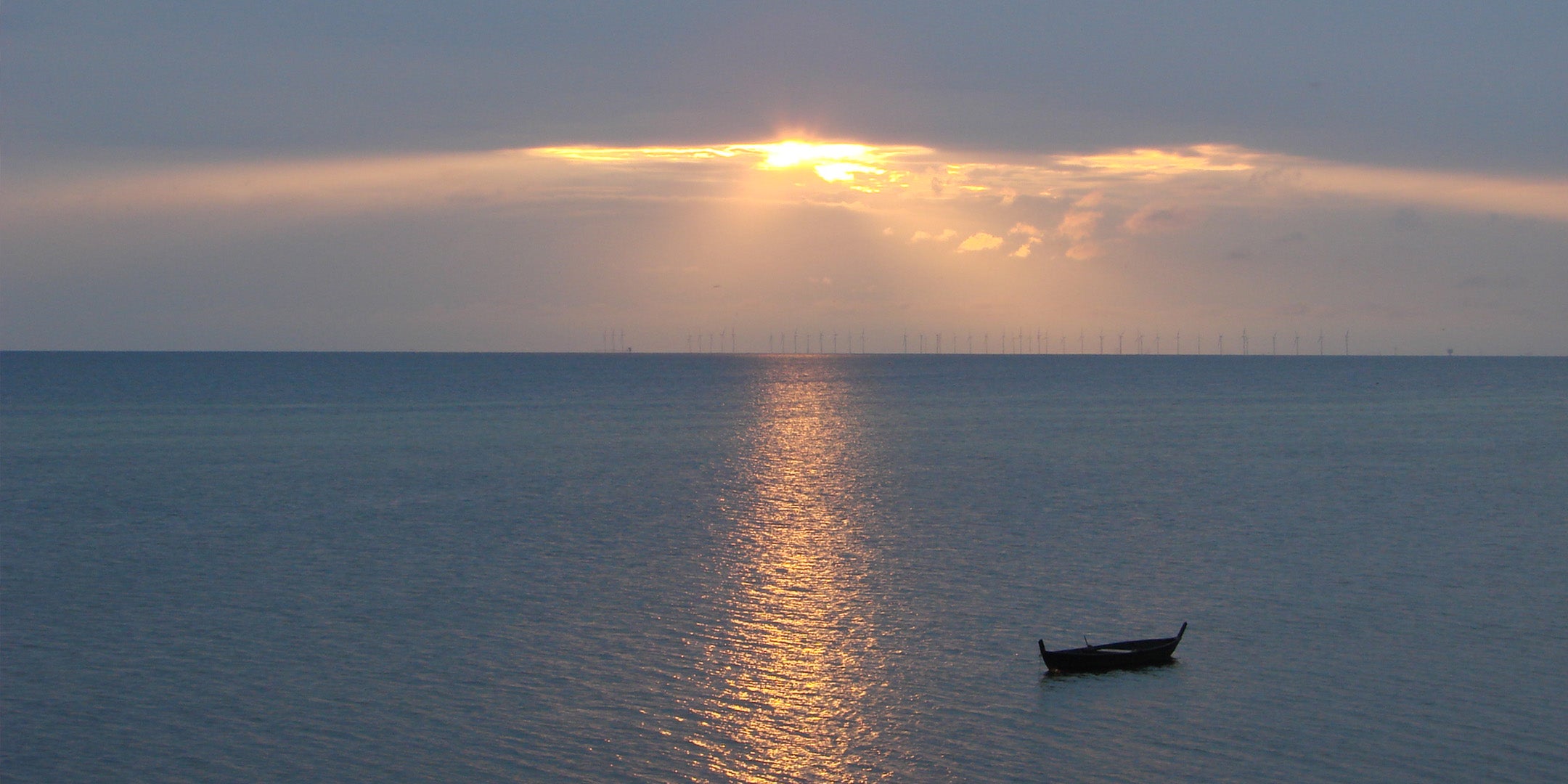Solnedgång över Öresund. Blått hav och mulen himmel möts. Molnen spricker upp och ljuset kommer igenom. Det speglas i en strimme på havet. I horisonten syns en lång rad vindkraftverk. En öppen träbåt ligger på vattnet