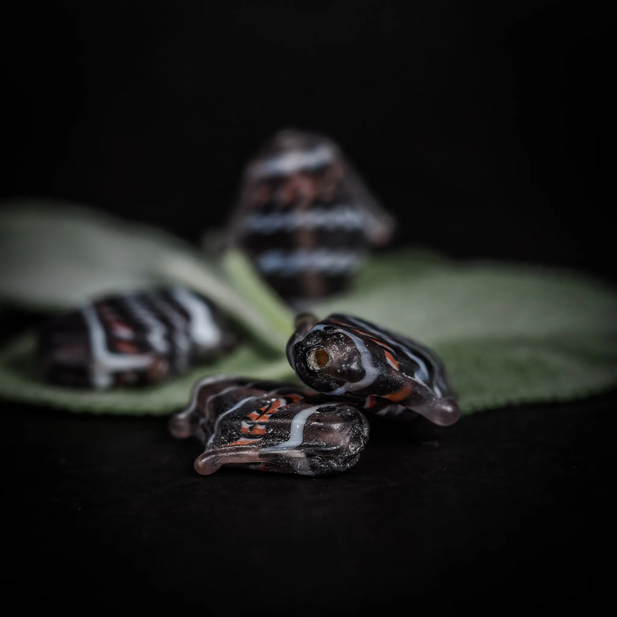 Handgjord glaspärla i form av fisk, grålila färg med rödvita mönster. Cirka 23x20x5mm med 1mm hål för tråd. Fotograferad på grön löv.