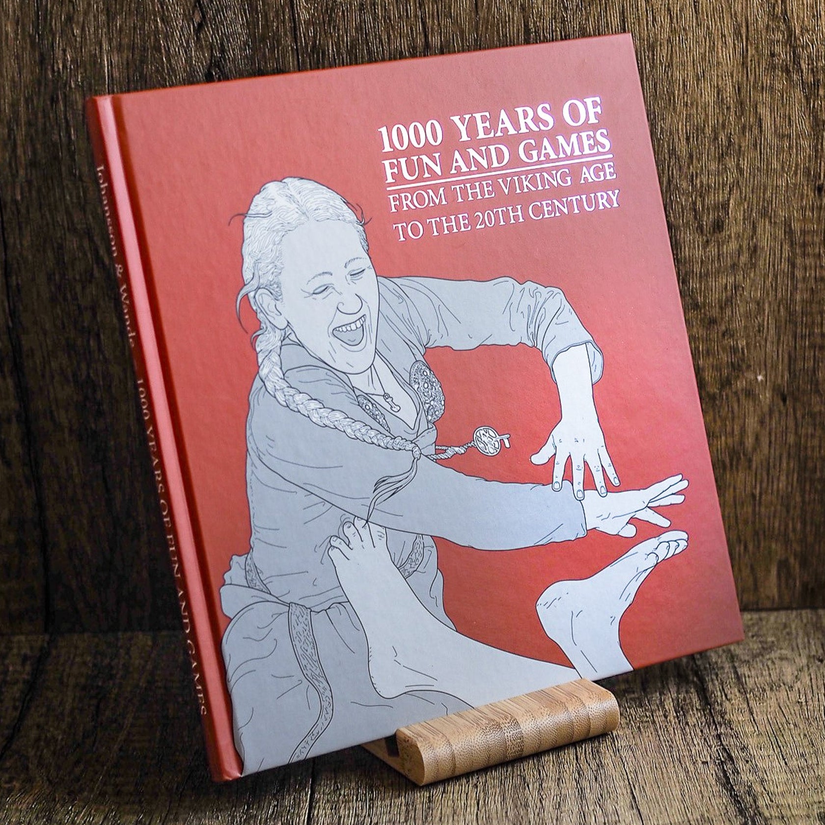 En inbunden bok med títeln '1000 Years of Fun and Games från vikingatiden till 20-talet' med terakotta-röd pärm och en illustrerad Viking-figur, stående på ett träverk.