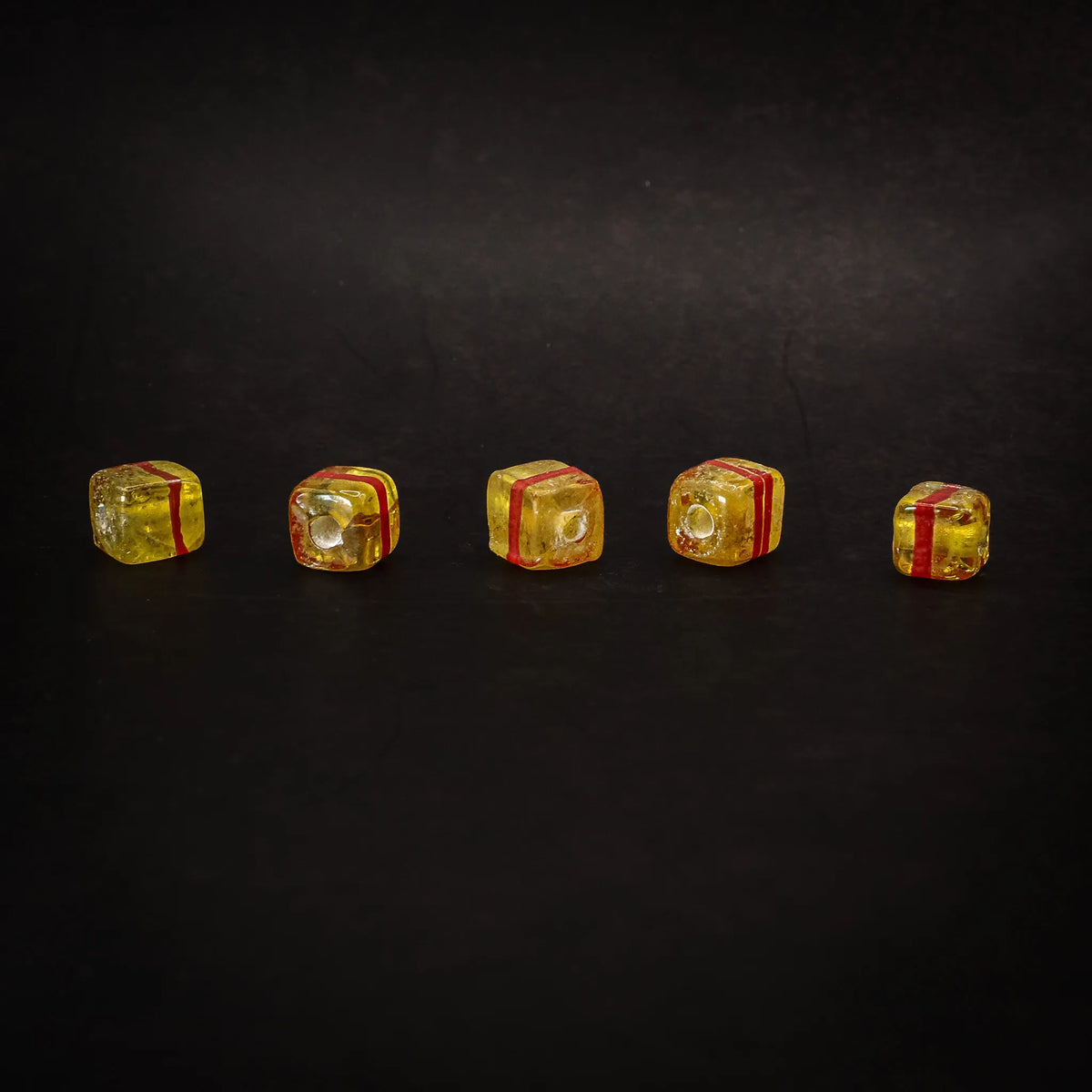 Fem gula kubiska glaspärlor i rad mot mörk bakgrund, varje med röd ränds, visar handgjord glasstruktur och 1mm håll