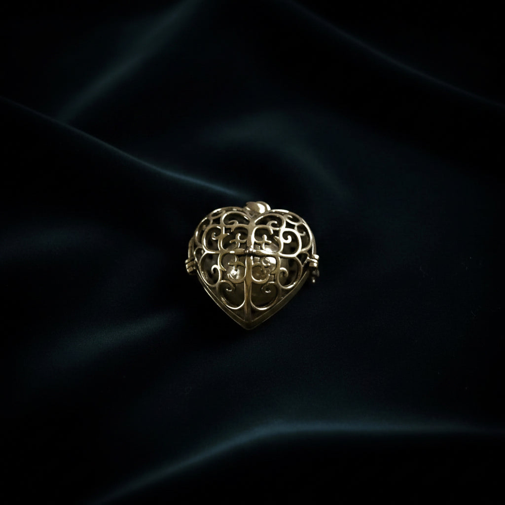 Mässing eller guldtonerad hjärtformad berlock med fint filigranmönster och genombruten ornamental design, fotograferad mot mörkblå tyg.