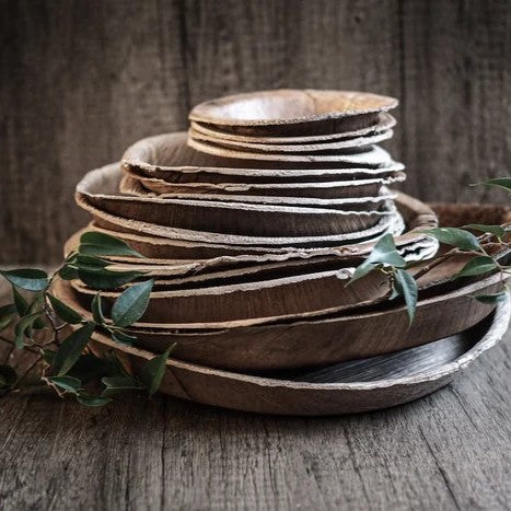Stapel av engångstallrikar i palmblad i naturlig färg mot rustik träbakgrund med gröna örter