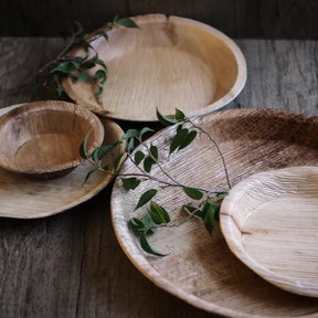 Engångstallrikar i palmblad i naturlig färg staplade på träyta med gröna örter för dekoration