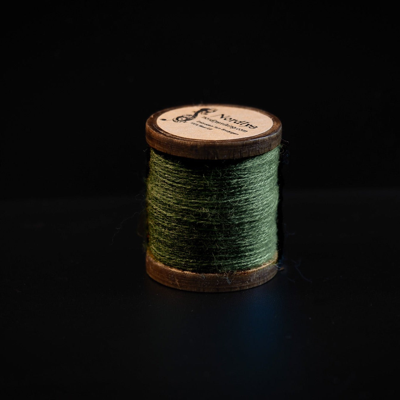 Grön brodyrtråd av 100% ull på träspole med Nordlys-märkning, fotograferad mot mörk bakgrund för att visa trådtextur och färg
