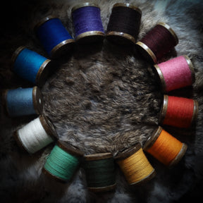 Tolv färgade brodyrtråd-spolar arrangerade i cirkel omkring djurhud på mörk bakgrund, visar traditionell nordisk textilkonstning